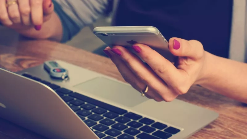 A woman is checking her mobile phone with a laptop on a desk in front of her.