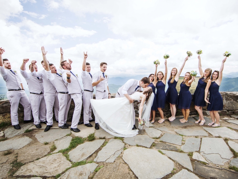 Bride and groom kissing while wedding party cheers at the top of the Blue Ridge Parkway.