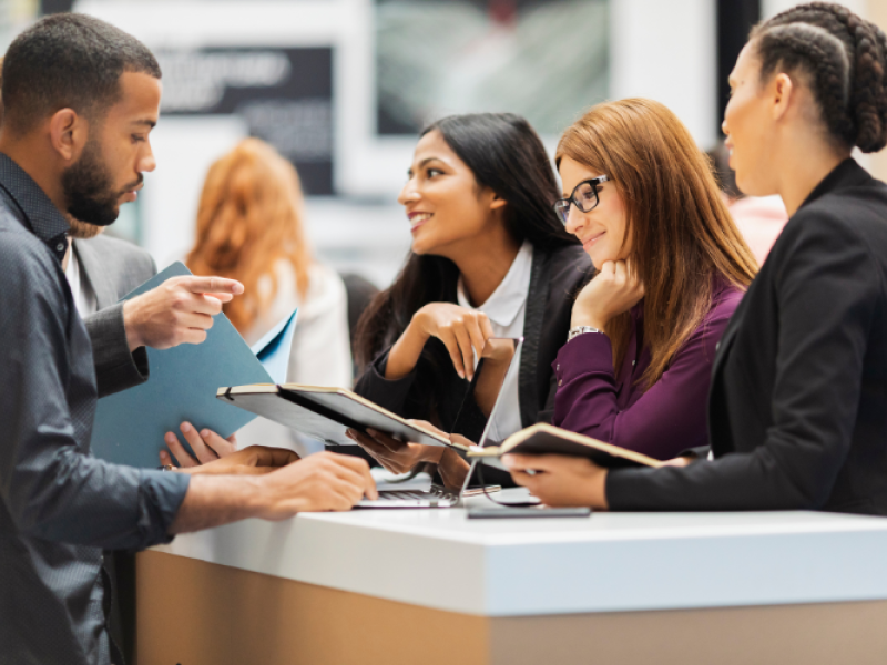 People in a business setting, discussing, with a laptop, folders, and a counter visible.