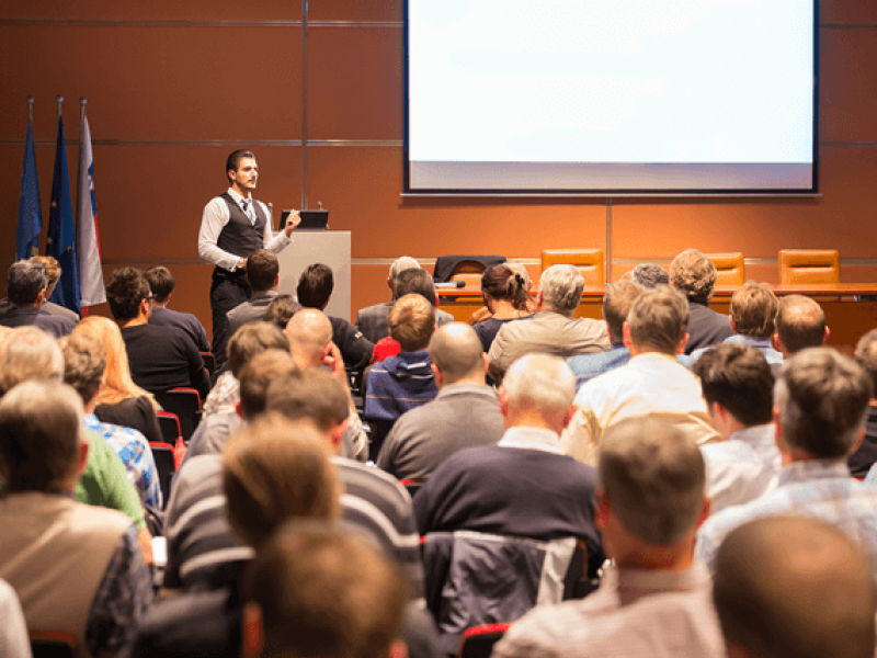 A man giving a presentation at a conference with an audience in front of him.