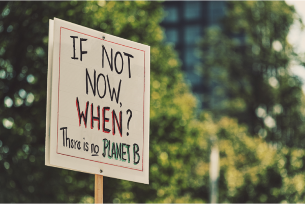 "Sign says 'If not now, when? There is no Planet B' in front of trees and architecture."