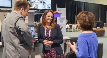 Two women and a man talk at an exhibit with a Connect Row banner and screens.