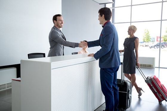 A man greets a guest at the front desk of a hotel while another guest walks by with luggage.