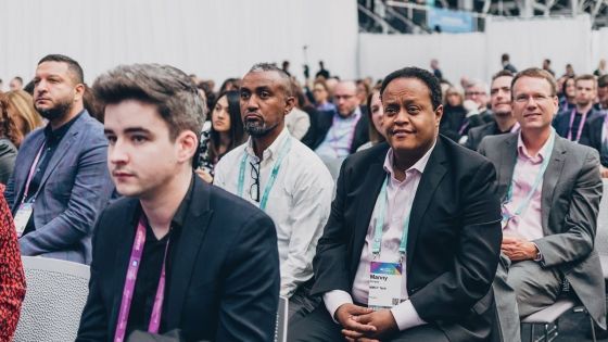 Several people are sitting in rows at an event, wearing name tags, and listening to a speaker.