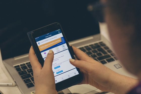 A woman is using her mobile phone to check an event guide while sitting in front of a laptop.