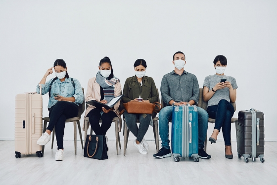 Six passengers wearing masks are sitting on chairs with luggage next to them, waiting for their flight.