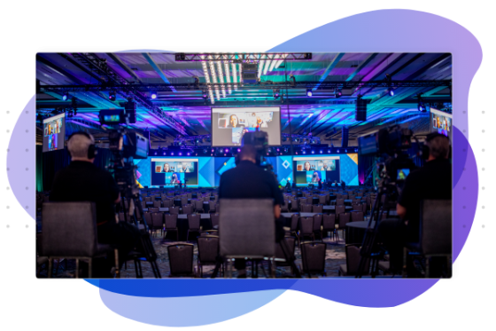 A group of people in a conference room with monitors, cameras, and a stage with purple lights.