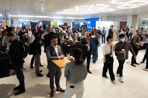 Group of people standing in a room with a desk, cup, handbag, TV, and lamps on the ceiling.