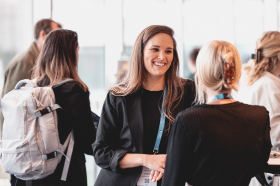 A smiling woman in a black jacket and blue lanyard converses with others in a conference setting.
