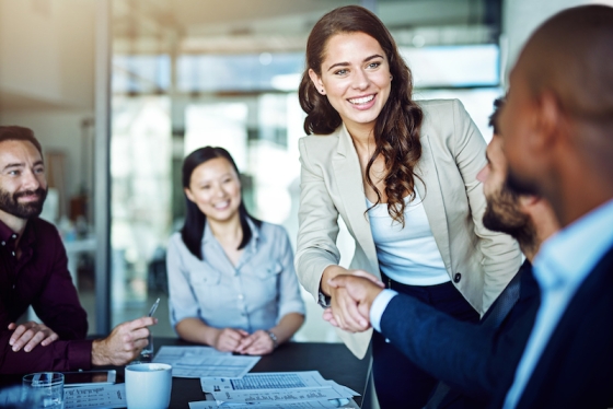 Four business people in a meeting shaking hands and smiling at each other with papers on the table.