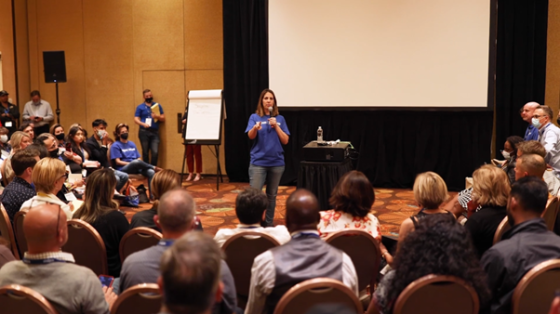 A woman stands on stage, speaking to a crowd in an auditorium with chairs, speakers, and a projector screen.