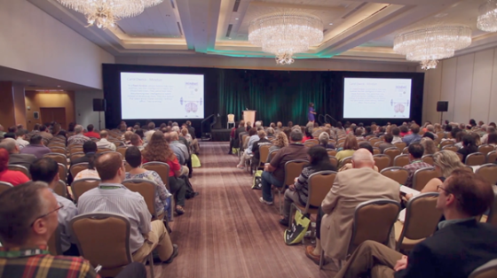People are sitting in chairs in a conference room with two big screens in front of them.