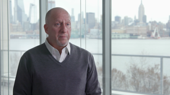 A man with a bald head standing in front of the glass window and wearing a gray sweater