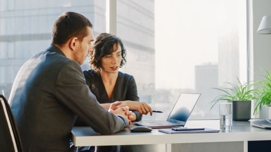 Two people sitting at a desk with a laptop and a glass of water on it.