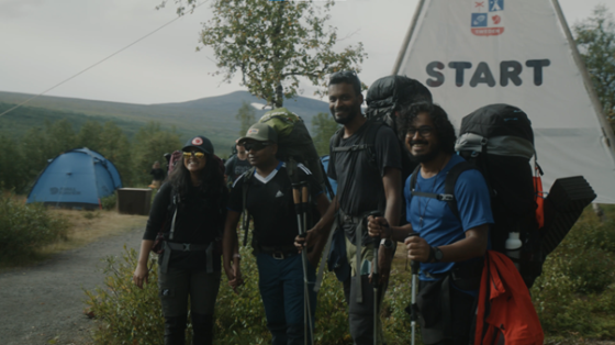 Five people in trekking gear pose in front of a START sign and a blue tent.