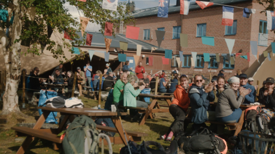 A group of people are sitting on benches with flags hanging above them in front of a building.