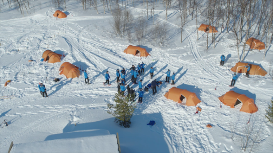 Several people are camping in the snow, with orange tents and trees in the distance.
