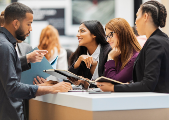 People in a business setting, discussing, with a laptop, folders, and a counter visible.