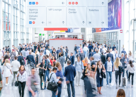 A large crowd of people at a convention with a large white sign hanging from the ceiling.