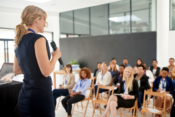 Woman speaking into microphone at conference with group of women sitting in chairs listening to her speak.