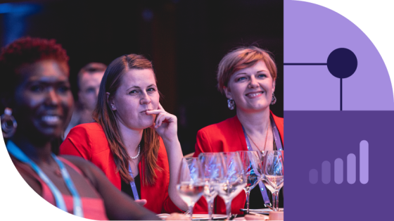 Three smiling women are sitting together at a table with wine glasses and a purple and black background.