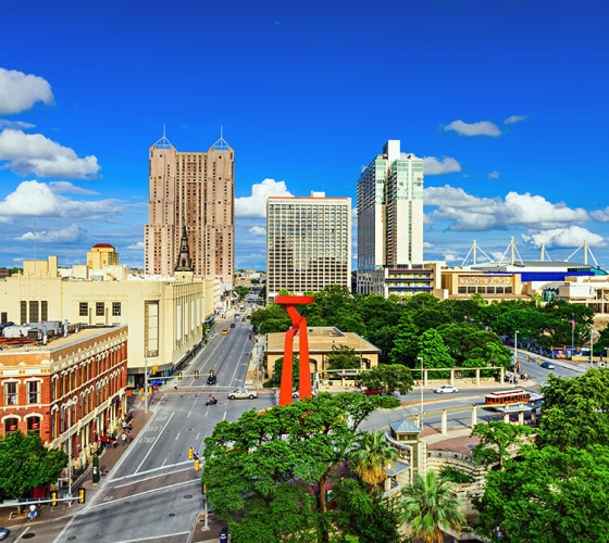 The cityscape of San Antonio with a red gate, a road, cars, trees, and tall buildings.