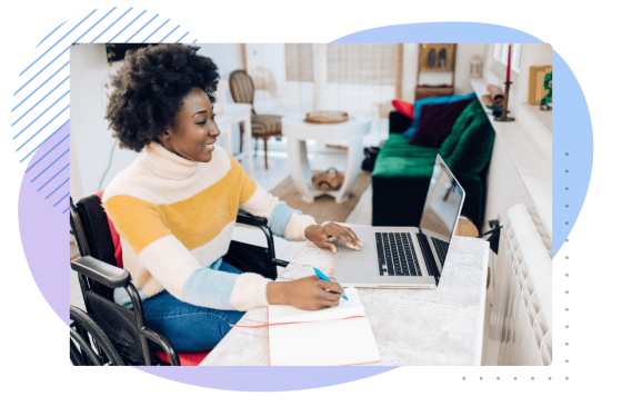 Woman in wheelchair writing on paper with pen and laptop on desk with living room in background.