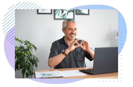 A man at his desk in his home office, he is smiling and making the sign for accessibility.