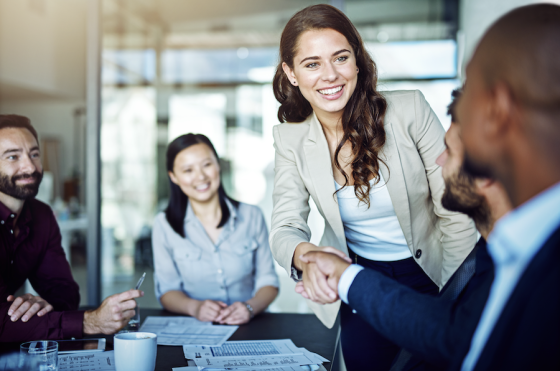 A group of four people in a meeting, shaking hands, and smiling in a bright office.