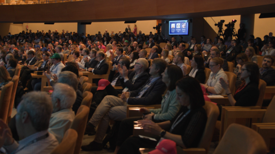 A crowd of people in an auditorium with a screen in the background, a few clapping their hands.