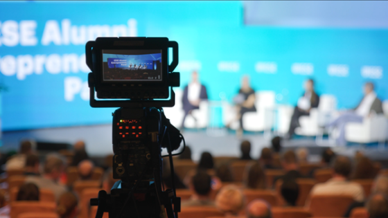 A video camera recording an event on a stage with a screen displaying text in the background.