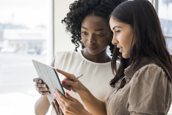 Two women discussing the tablet with each other in front of the glass wall in the office.