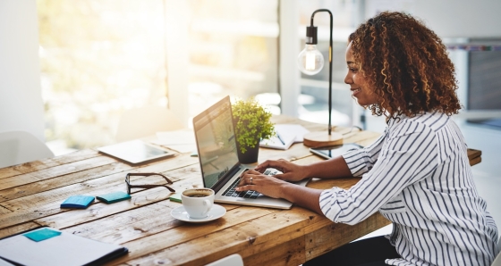 Woman wearing striped shirt working on laptop and drinking coffee on wooden table in a modern office.