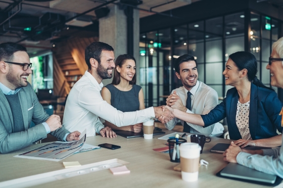 A group of people in an office shaking hands around a table with coffee and documents.