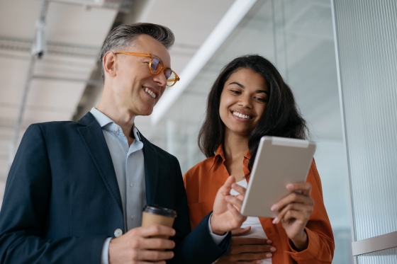Two people smiling while looking at a tablet in a modern, glass-paneled office setting.