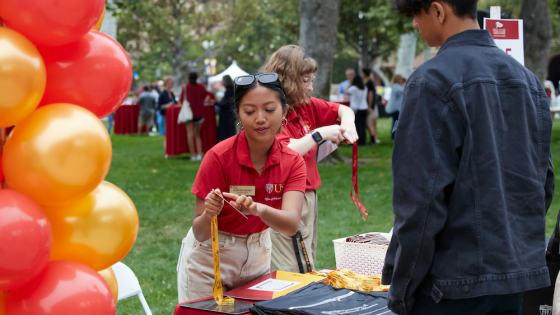 Two women and a man stand near balloons, one woman holding a badge and a pen.