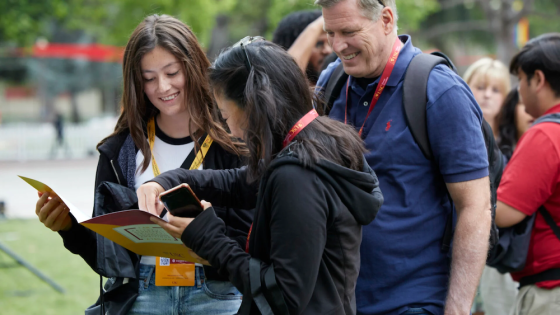 Two women and a man with lanyards around their necks look at a paper and a phone.