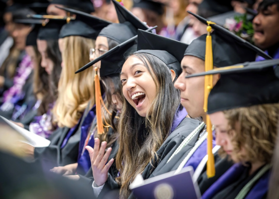 Graduates at Portland State University are dressed in robes and caps and are clapping while sitting in chairs.