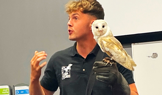 A man in a black polo shirt is holding a barn owl while talking in front of a whiteboard.