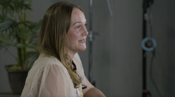 A woman with blonde hair and freckles sits in front of a potted plant and gray wall.