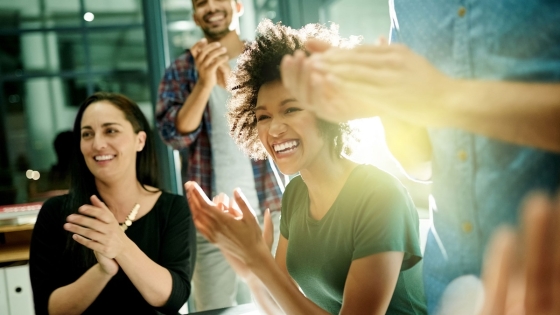 Smiling and clapping woman at the center of a group of people, close-up, indoors, warm lighting.