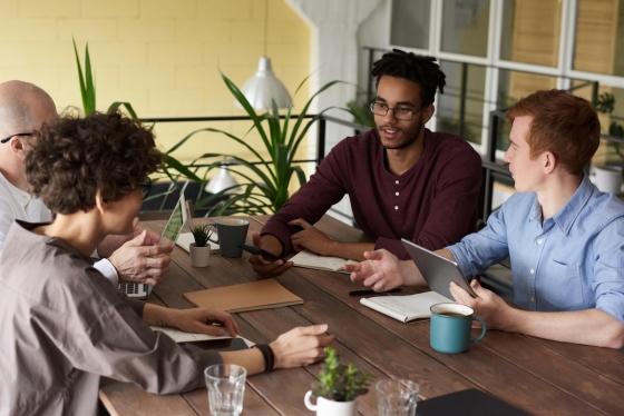 Four people sitting around a table with laptops and a plant on it and a railing behind them.