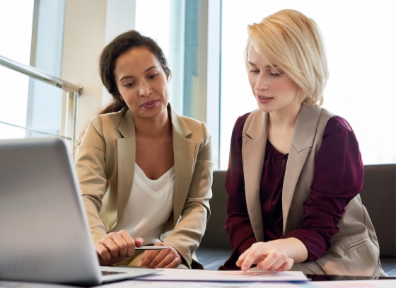 Two women in business attire look at documents on a laptop in a modern office environment.