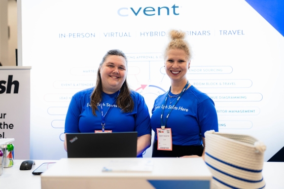Two women stand behind a table with a laptop, smiling at the camera at Cvent INPEX.