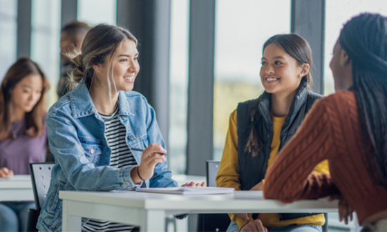 Three women are seated at a table, talking to one another in a school setting.