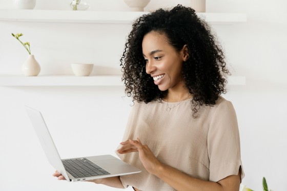 A woman is smiling while holding a laptop in front of a shelf with some vases.