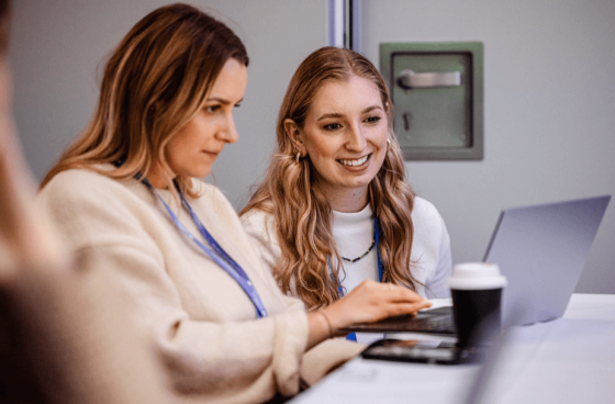 Two women are working together on a laptop, smiling, and enjoying a coffee.