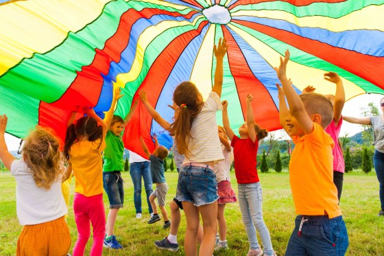 Kids having fun outside with a colourful parachute with adults nearby on a sunny day.
