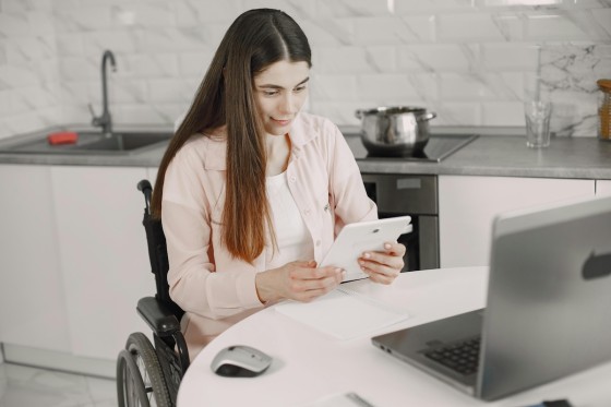 Woman in wheelchair using tablet and laptop in a kitchen with white tiles and modern appliances.