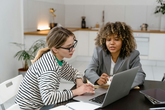 Two women are sitting in front of a laptop on a table in a modern kitchen.